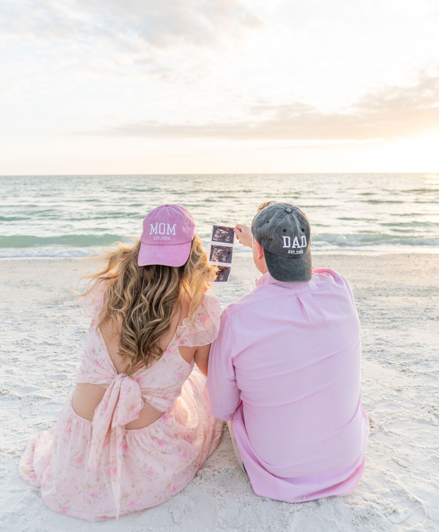 Couple sitting on beach wearing Mom and Dad hats, holding ultrasound photo, pregnancy announcement at sunset.