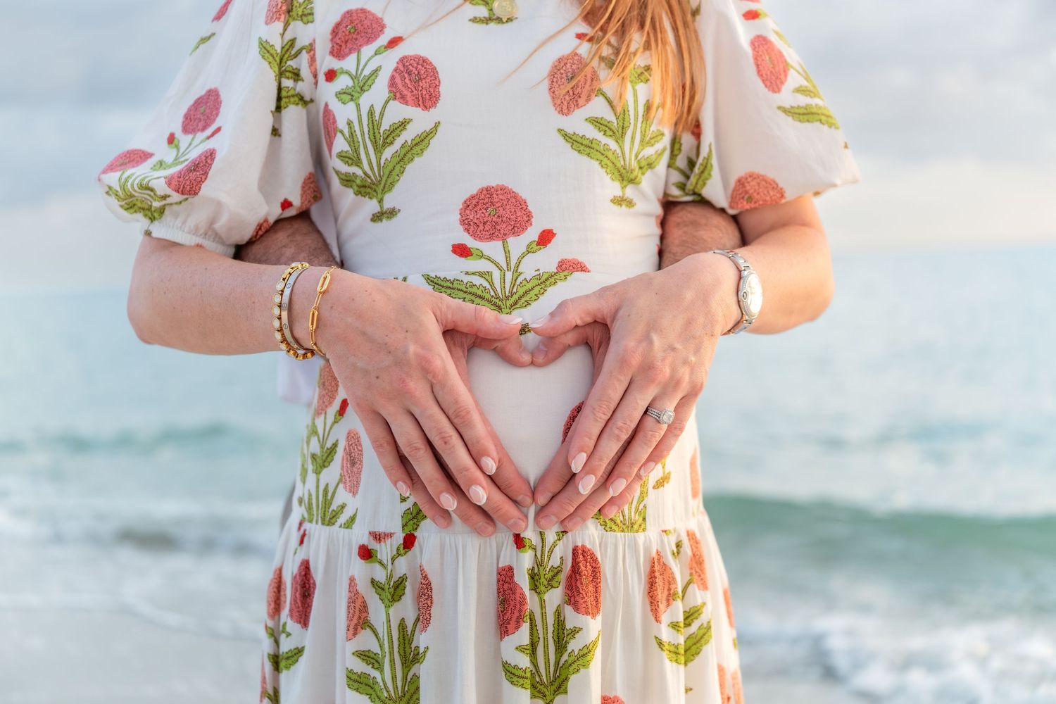 Pregnant woman in floral dress making heart shape with hands on baby bump at beach.
