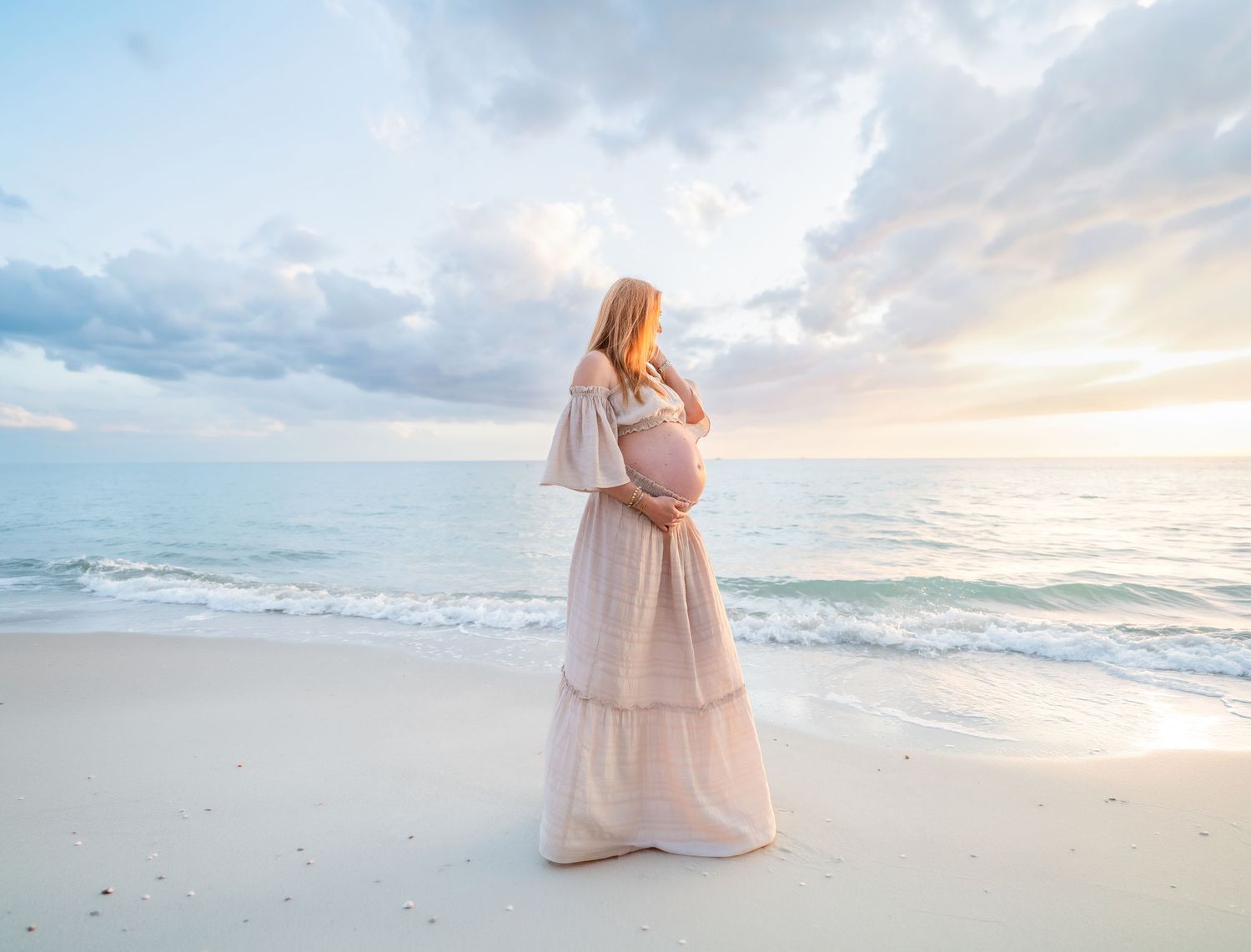 Pregnant woman in white dress standing on beach at sunset, cradling her baby bump while gazing at the ocean.