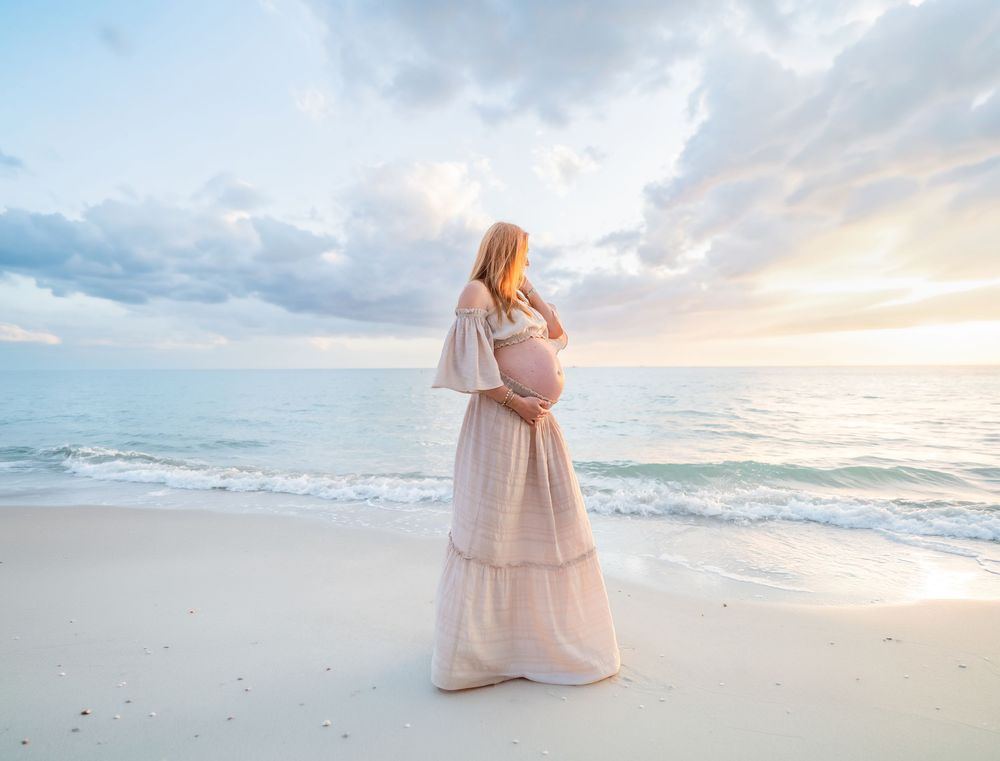 Pregnant woman in white dress standing on beach at sunset, cradling her baby bump while gazing at the ocean.