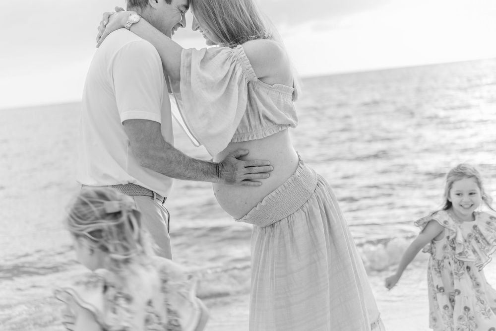 Black and white beach maternity photo of couple embracing with two young girls playing around them near the ocean.
