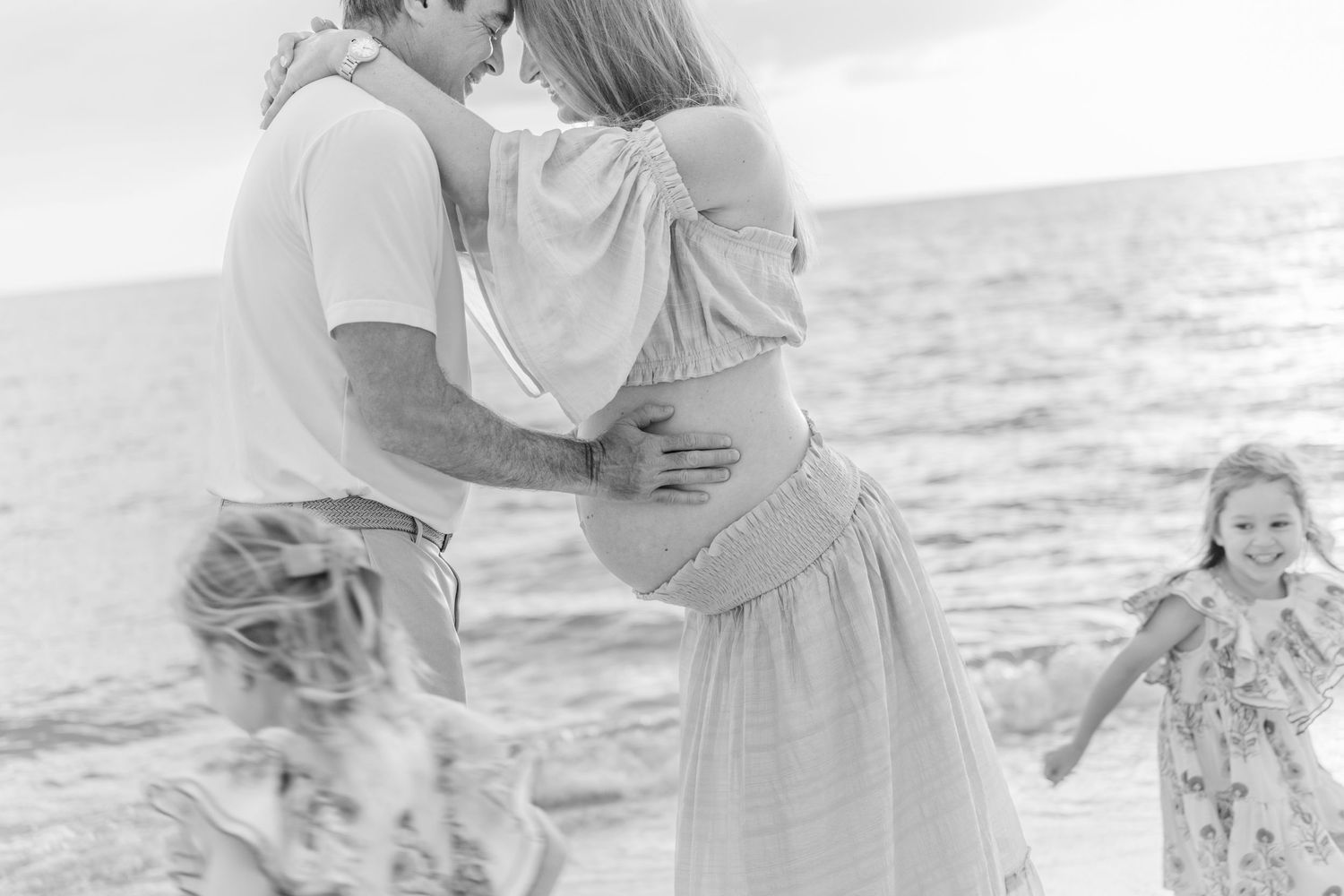 Black and white beach maternity photo of couple embracing with two young girls playing around them near the ocean.