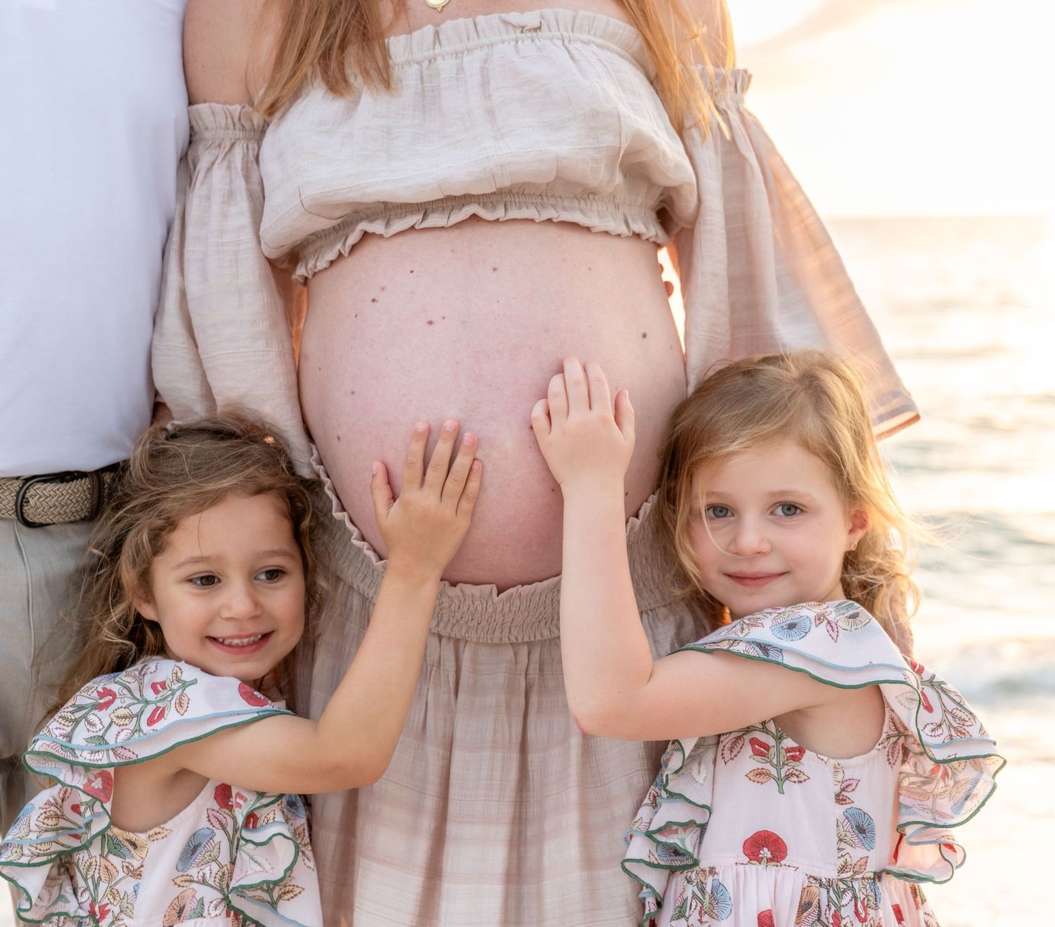 Two young girls touching pregnant mothers bare belly during a beach maternity photo session.