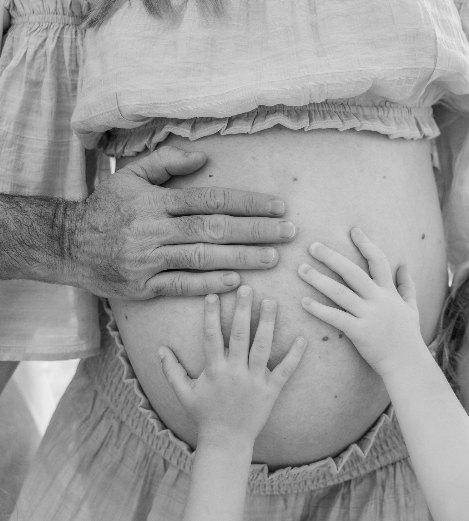 Black and white photo of pregnant woman's belly with adult and child hands gently resting on it.