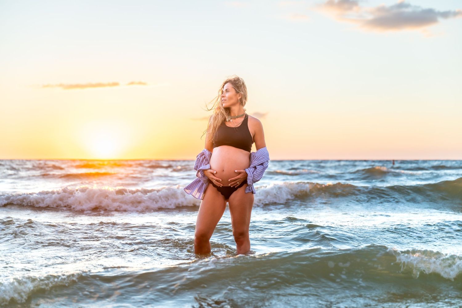 A person in a black swimsuit stands in the ocean waves during a vibrant golden sunset.