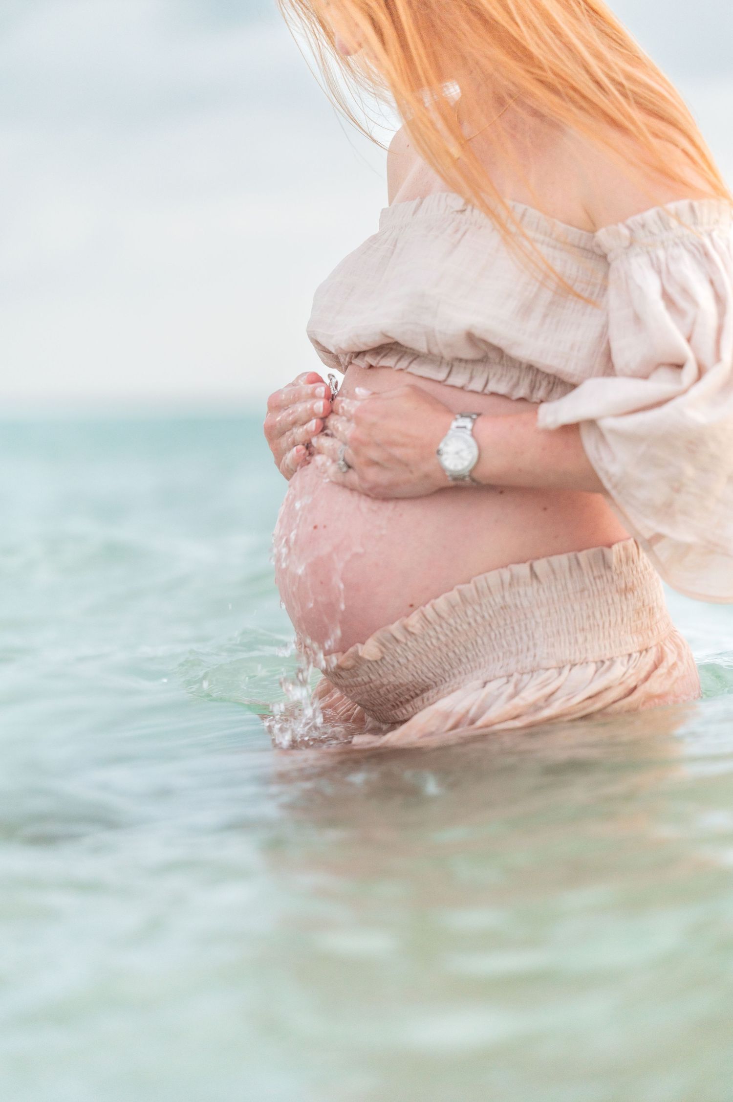 Pregnant woman in beige off-shoulder dress standing in ocean water, cradling her baby bump.