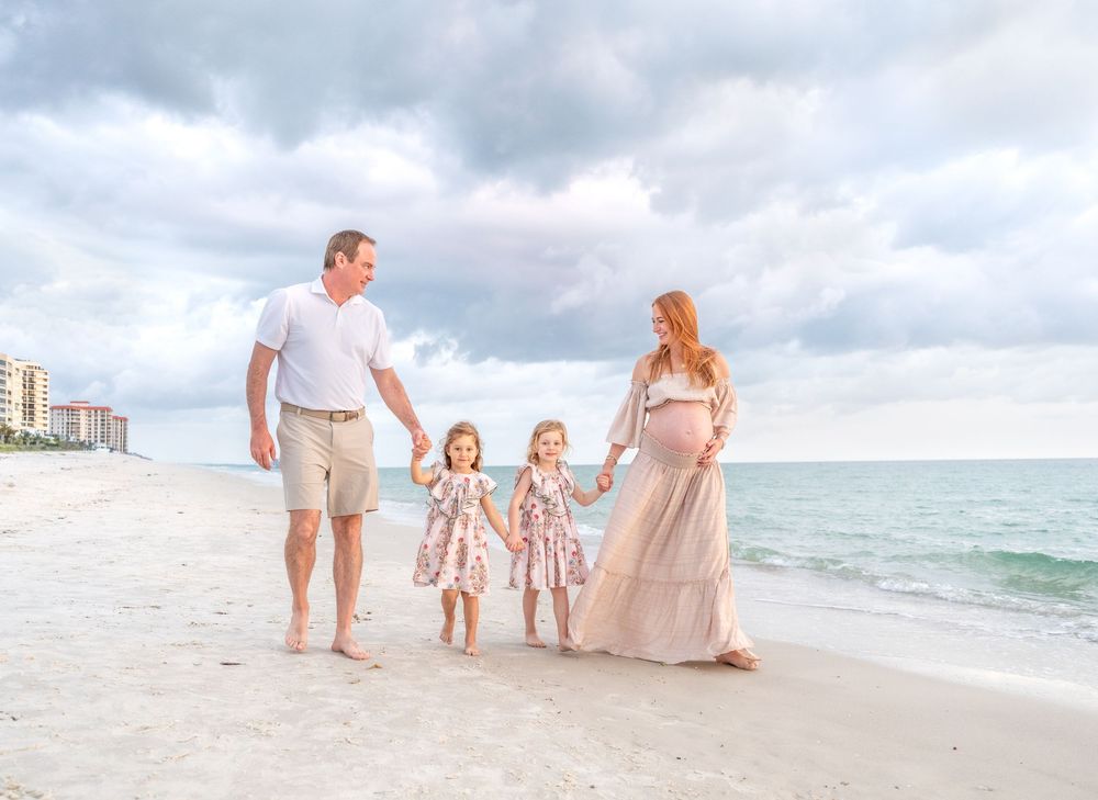 Pregnant woman and man walk on beach with two young girls in matching floral dresses under cloudy skies.
