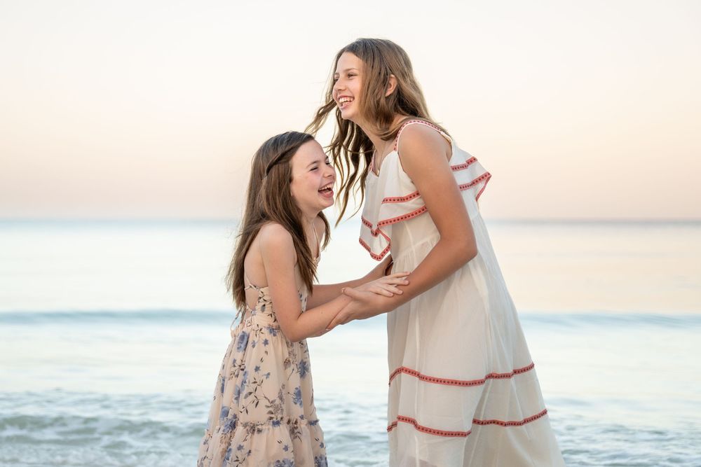 Two girls laughing and embracing on a beach at sunset, wearing floral and white dresses.