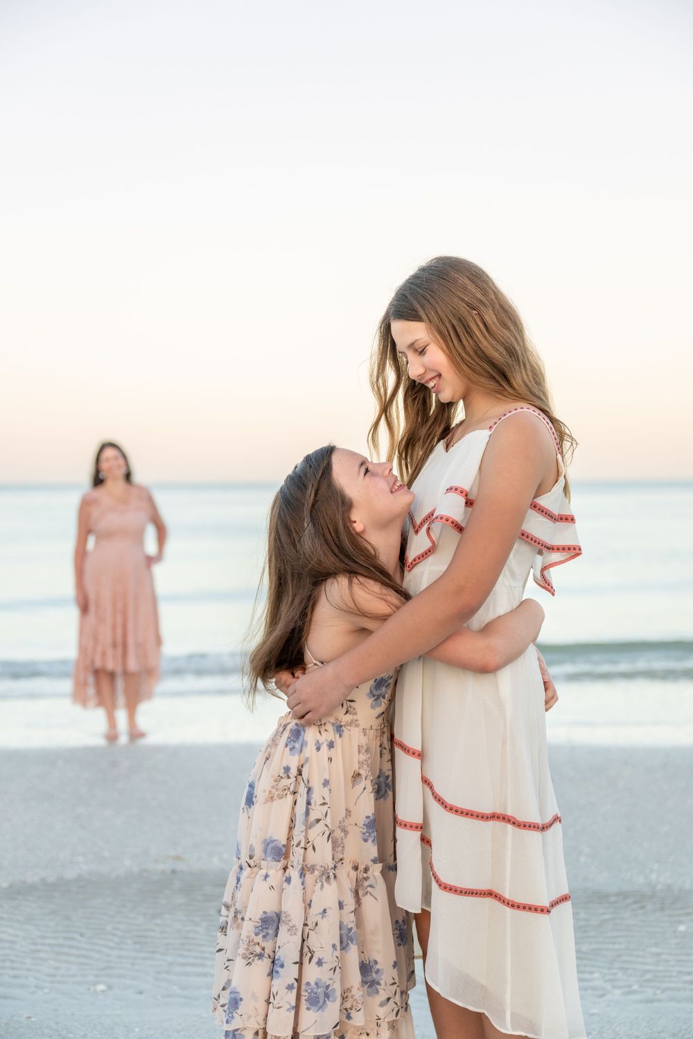 Two girls in floral dresses embrace on a beach at sunset, smiling at each other, with a woman standing in the background.