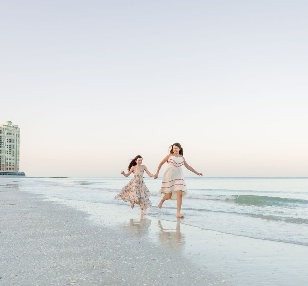 Two girls in floral dresses hold hands running along a sunny beach with calm waves and a building in the background.