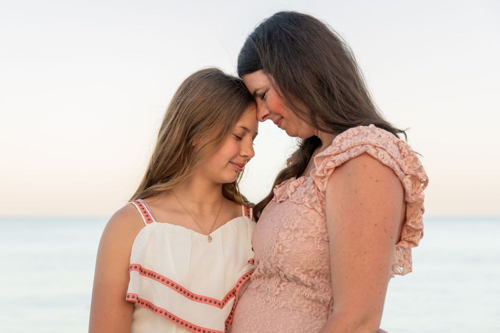Mother and daughter share a tender forehead-to-forehead moment on a beach at sunset in matching pink dresses.