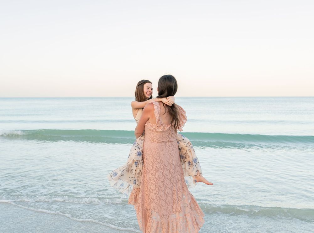 A woman in a pink lace dress lifts a child on a serene beach at dusk with calm ocean waves behind them.