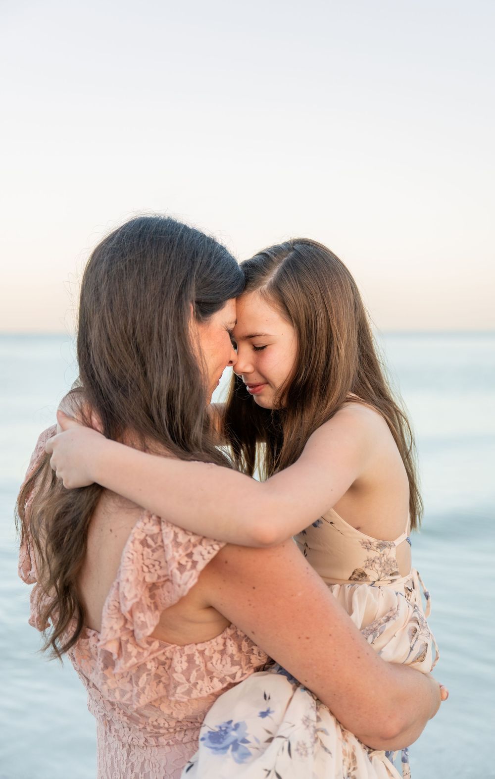 Two girls with long brown hair share a tender forehead-touching hug on a beach at sunset in floral dresses.