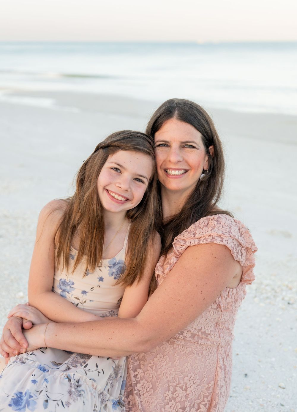 A woman and young girl smile together on a white sand beach, wearing floral dresses in a warm, candid portrait.