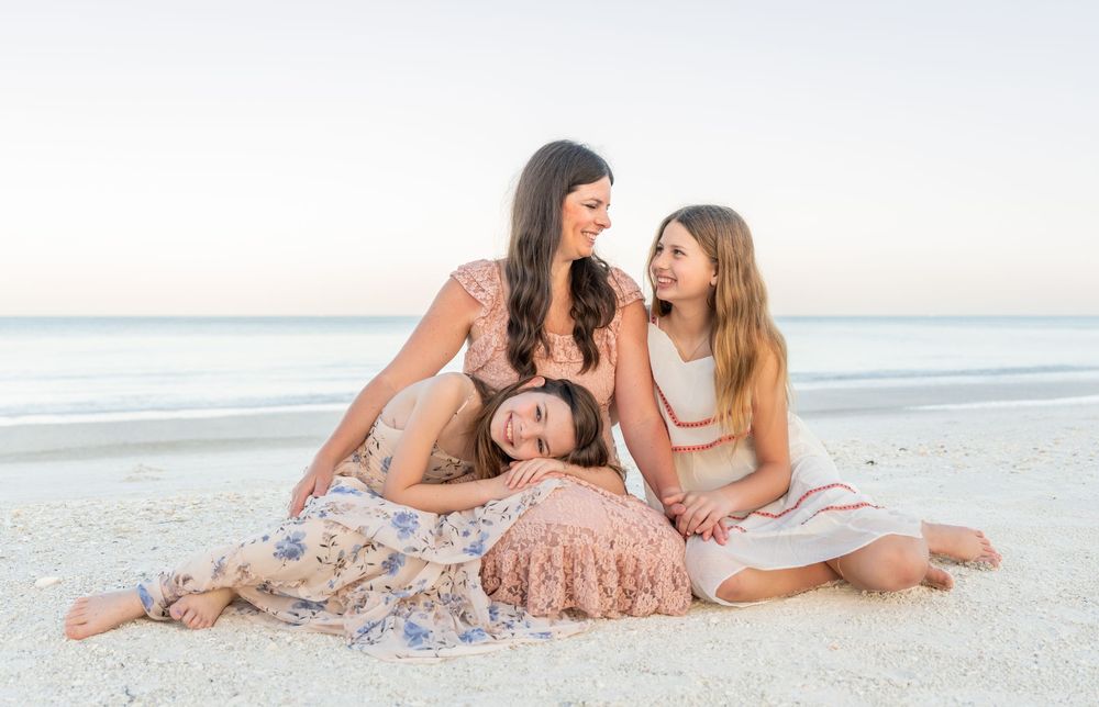 Three women smiling and relaxing together on a sandy beach at sunset in floral and pastel dresses.