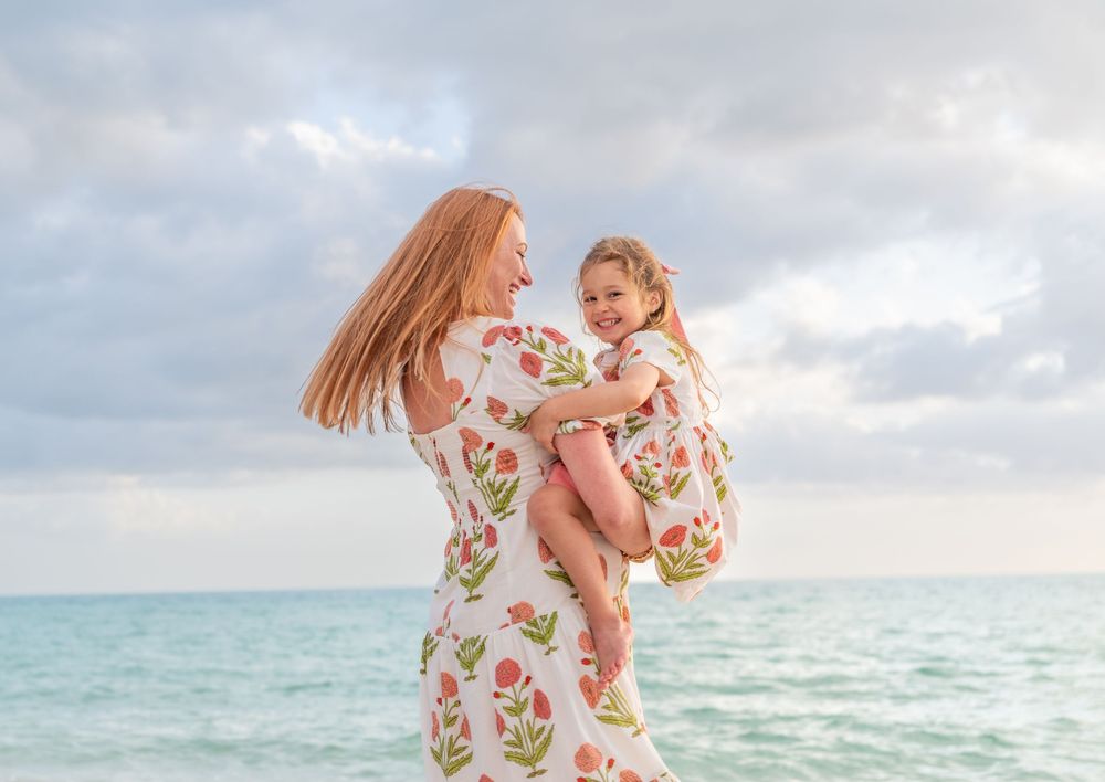 A mother in a floral dress lifts her smiling daughter on a beach, both wearing matching outfits by the ocean.