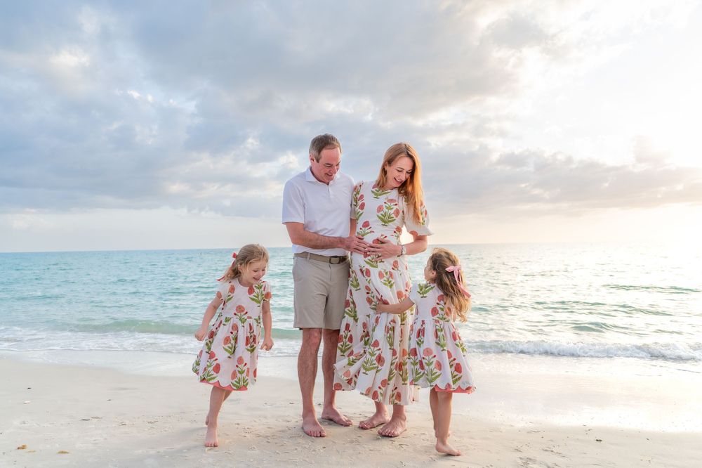 Pregnant woman with husband and two young daughters in matching floral dresses on a beach at sunset.