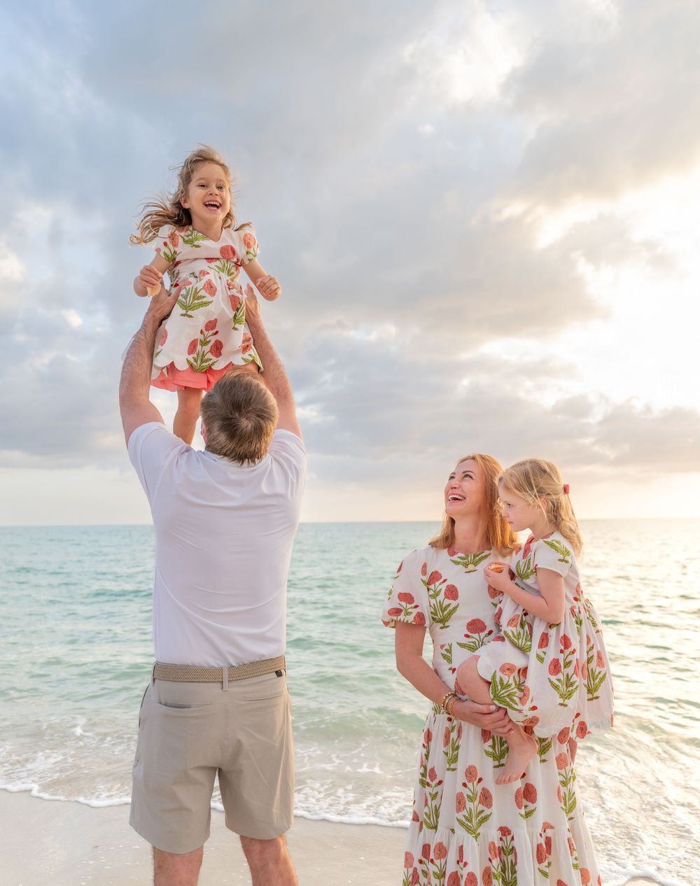 Family beach photo with dad lifting daughter in floral dress as pregnant mom holds child at sunset.