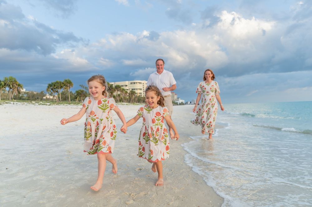 Family of four walking on white sand beach, two girls in floral dresses holding hands, parents behind them, cloudy sky.
