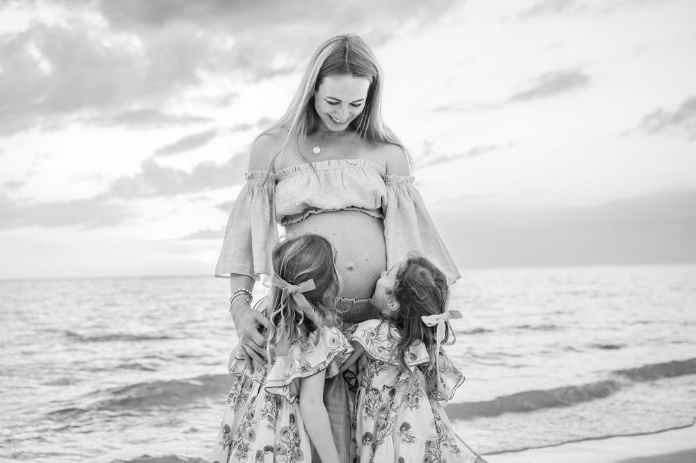 Pregnant woman on beach with two young daughters hugging her baby bump in black and white maternity photo.