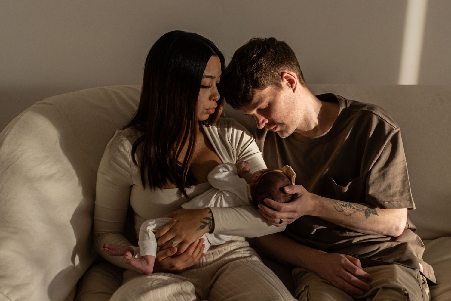 A young couple holds their newborn baby girl on a couch in studio with natural light coming in from the right