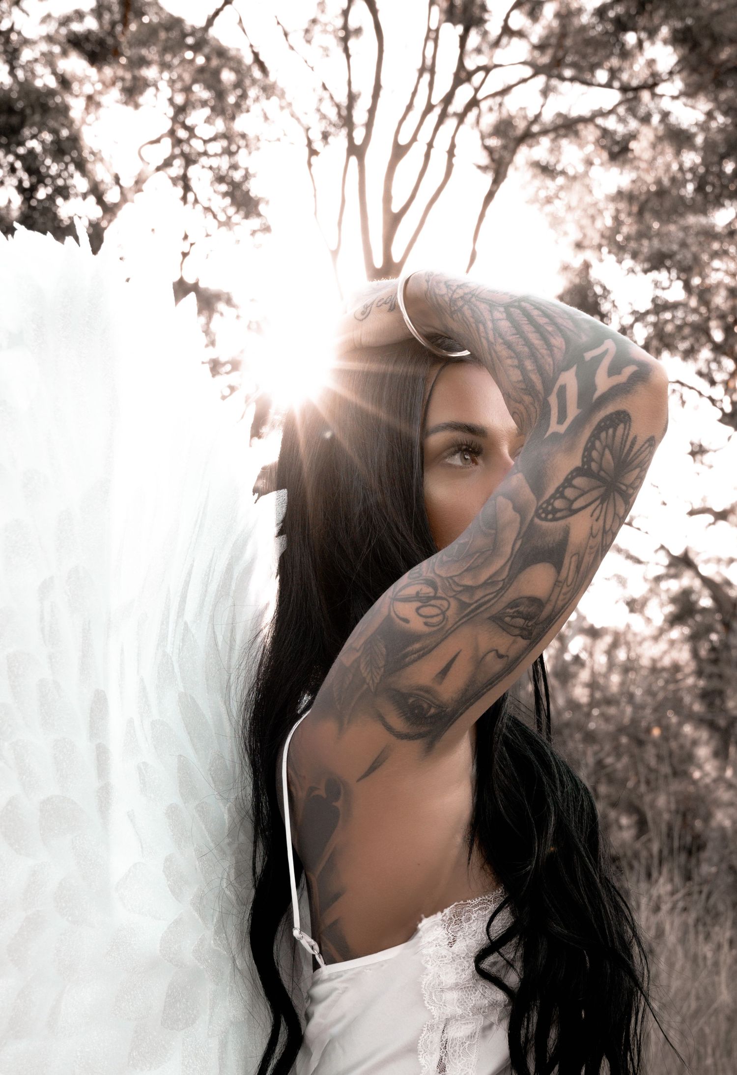 Artistic black and white portrait of a person with dark hair and arm tattoos looking away from the camera.