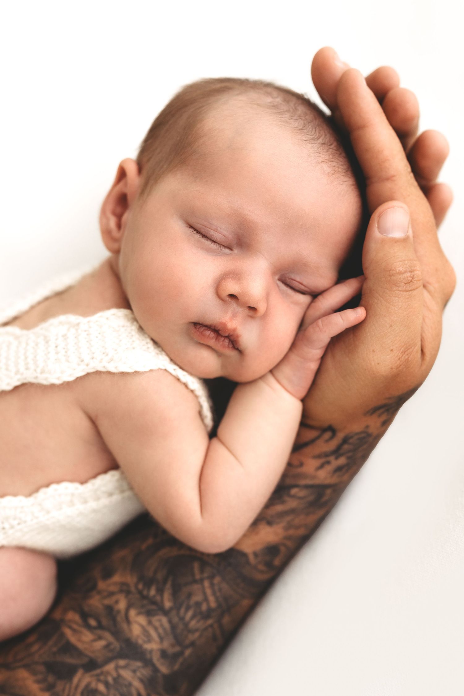 Sleeping newborn baby resting on tattooed fathers hand, wearing white knit outfit on white background.