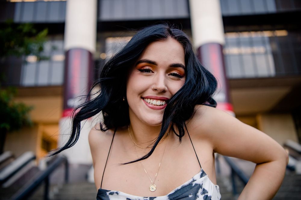 A happy person with black wavy hair in a floral summer dress smiles outside a building with columns.