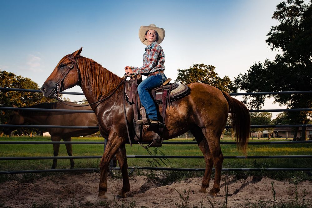 Someone in a straw hat and jeans sits atop a brown horse in a sunny outdoor arena at golden hour.