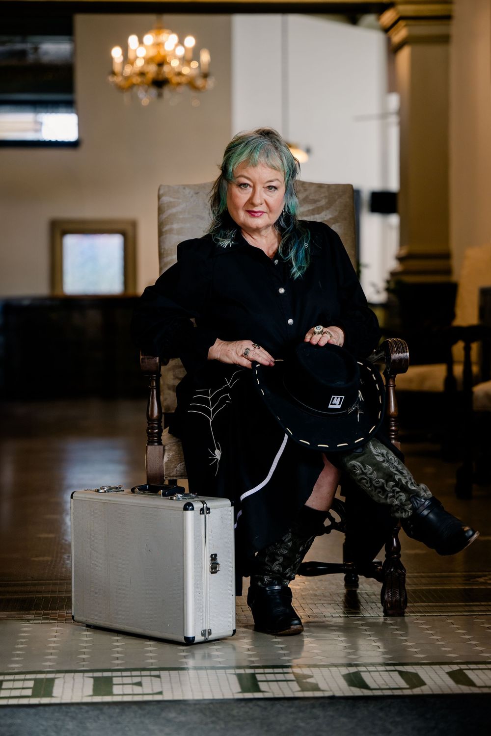 Someone sits on a tiled floor wearing black clothing while posing with a metallic silver briefcase in an elegant room.