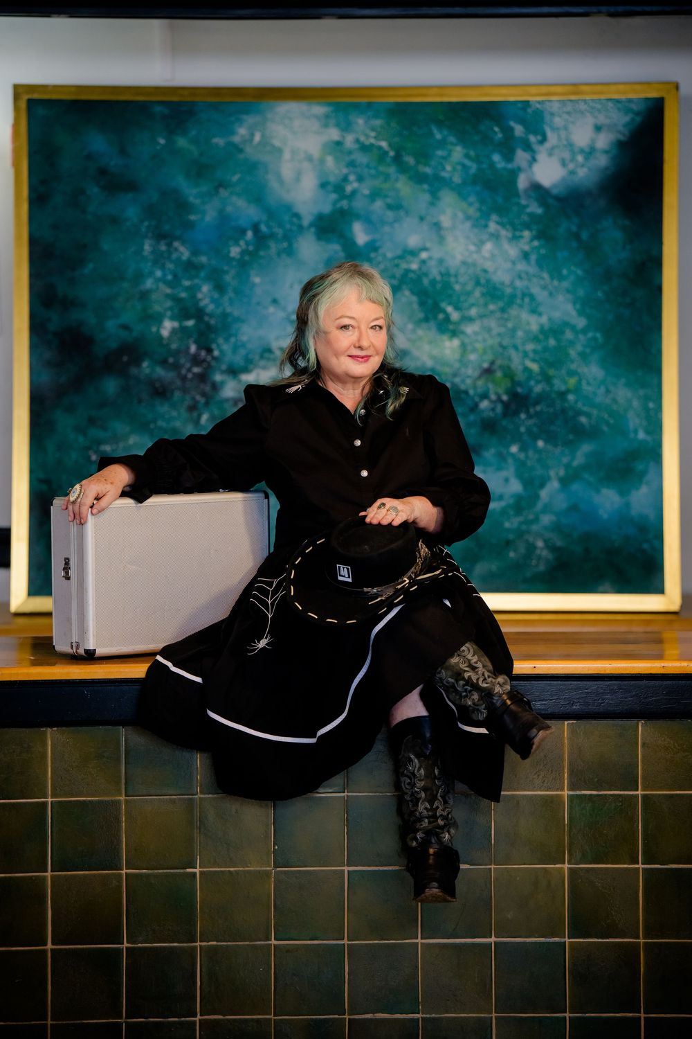 A person in a black dress and boots sits on a ledge against a dreamy turquoise backdrop with dark tiled wall.