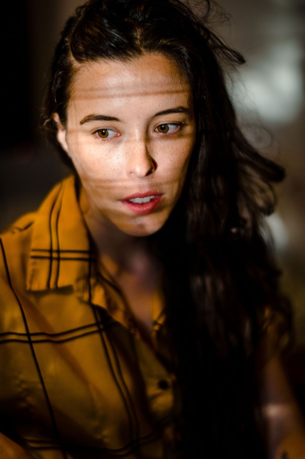 Portrait with soft lighting showcasing long dark hair and a mustard yellow plaid shirt in moody ambiance.