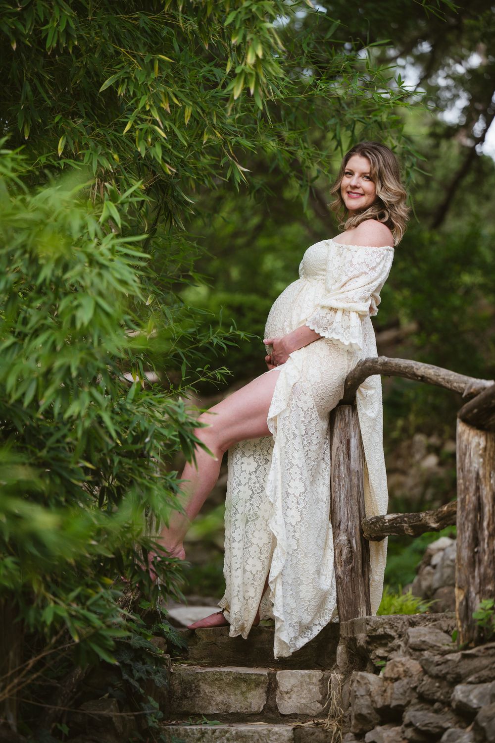 A romantic maternity photo shows an expectant mother in a lace dress sitting on rustic stone steps surrounded by greenery.