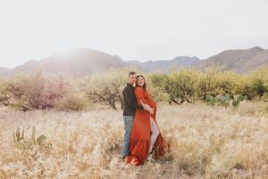Desert maternity photoshoot with flowing red dress against mountain backdrop and desert brush.
