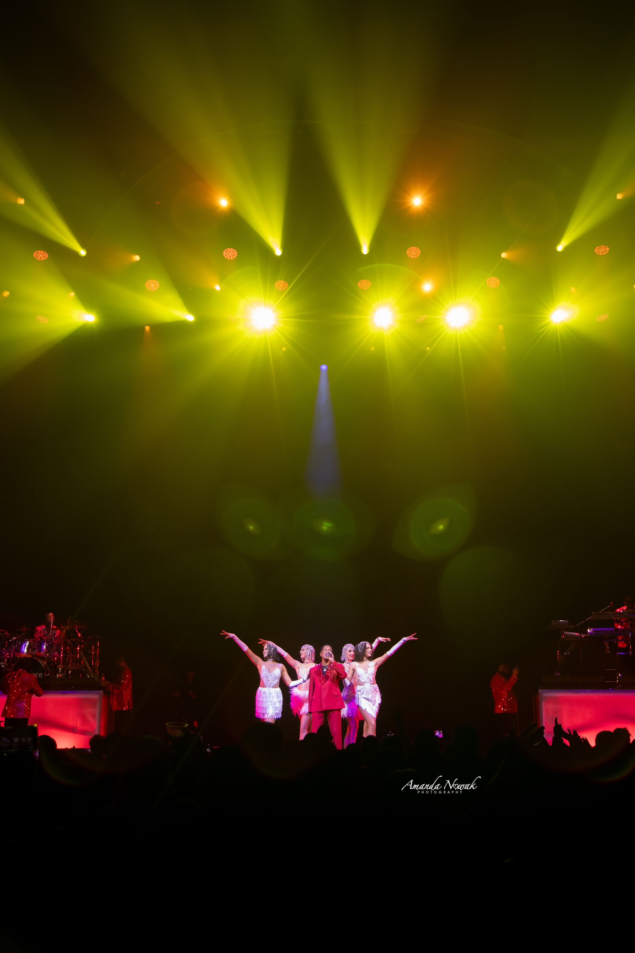 Four performers in colorful costumes on stage under dramatic green concert lighting.
