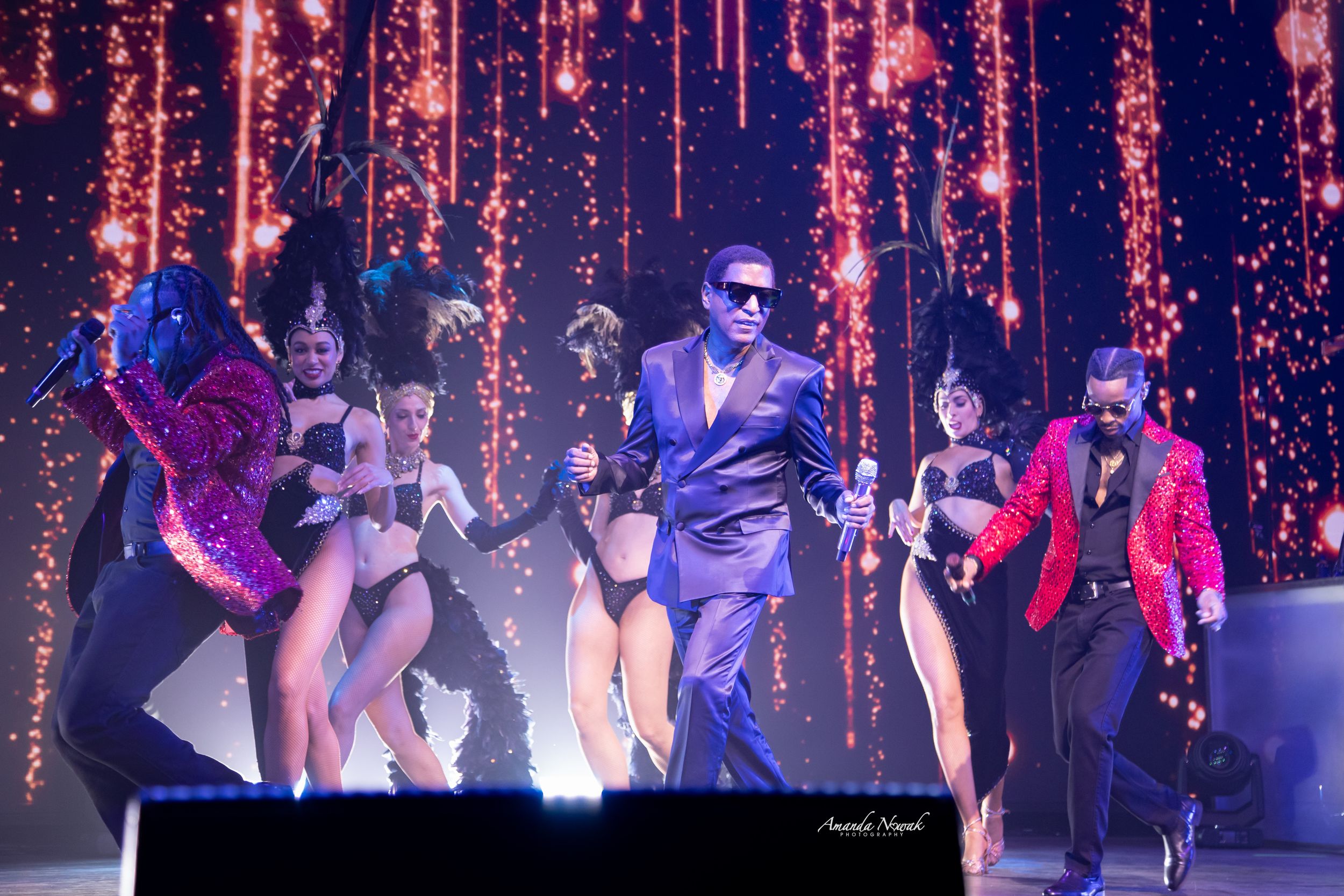 Male performer in blue suit dancing with backup dancers under red sparkle lights.