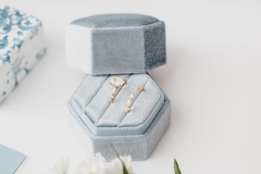 Two delicate gold rings with pearl and diamond details displayed in a grey velvet hexagonal ring box.