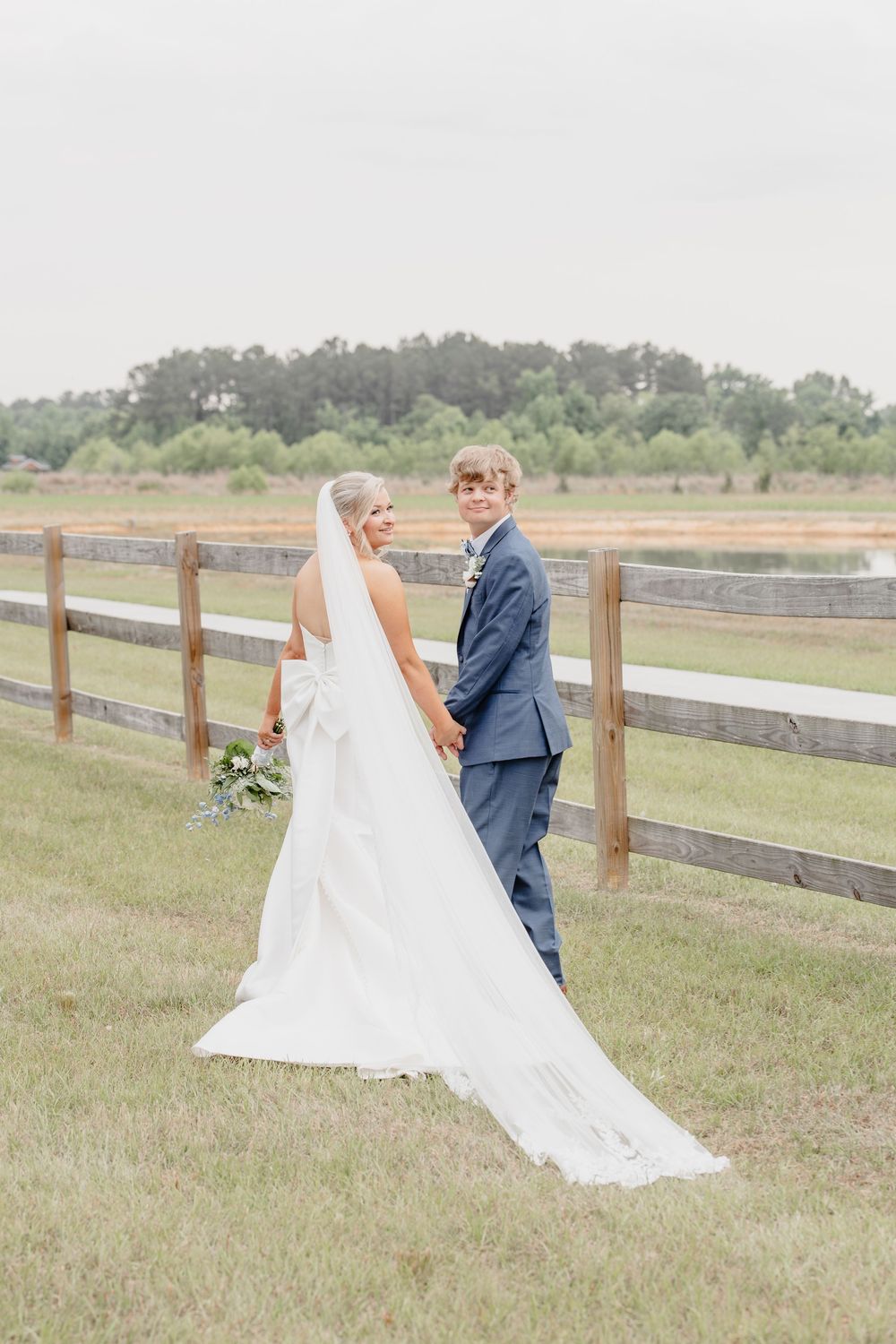 Bride and groom holding hands by a wooden fence on their wedding day, bride wearing a long white gown with veil.