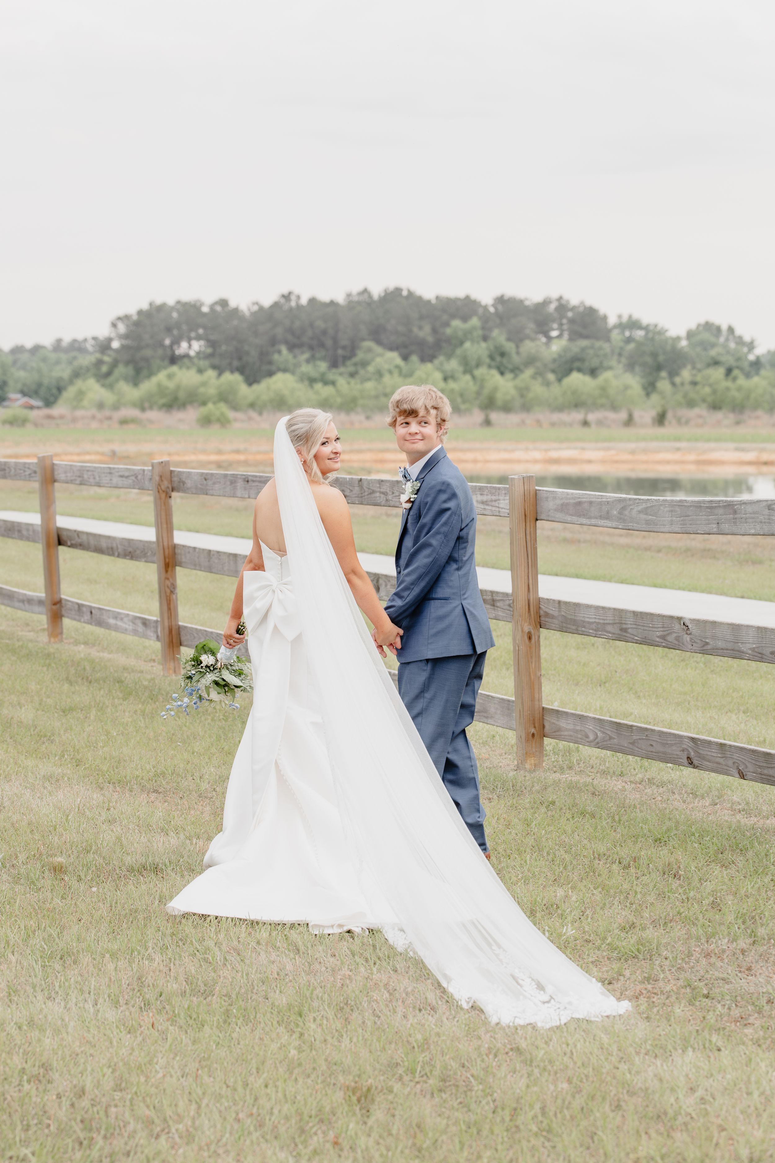Bride and groom holding hands by a wooden fence on their wedding day, bride wearing a long white gown with veil.