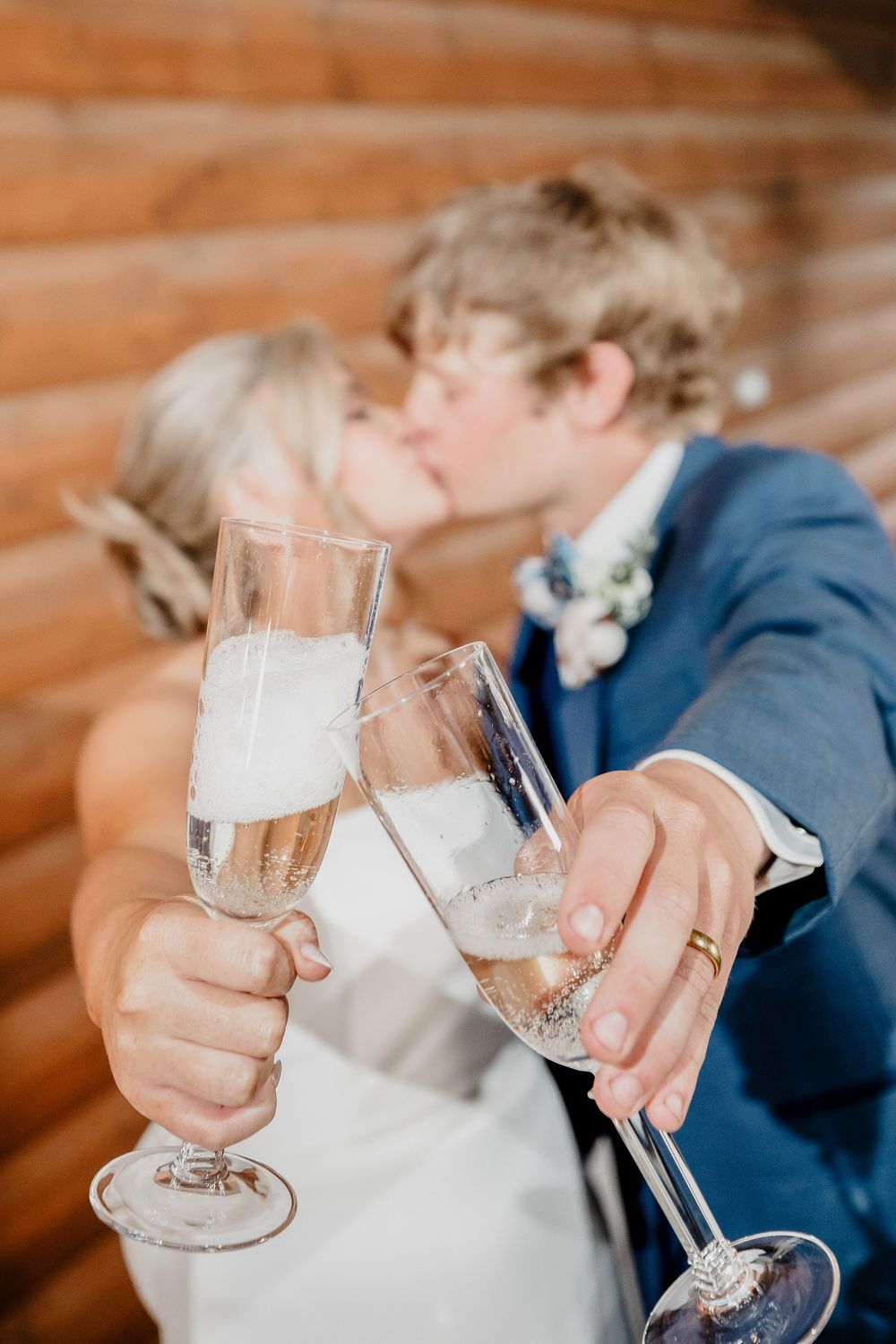 Newlywed couple kissing while clinking champagne flutes, wearing wedding attire near a rustic log wall.