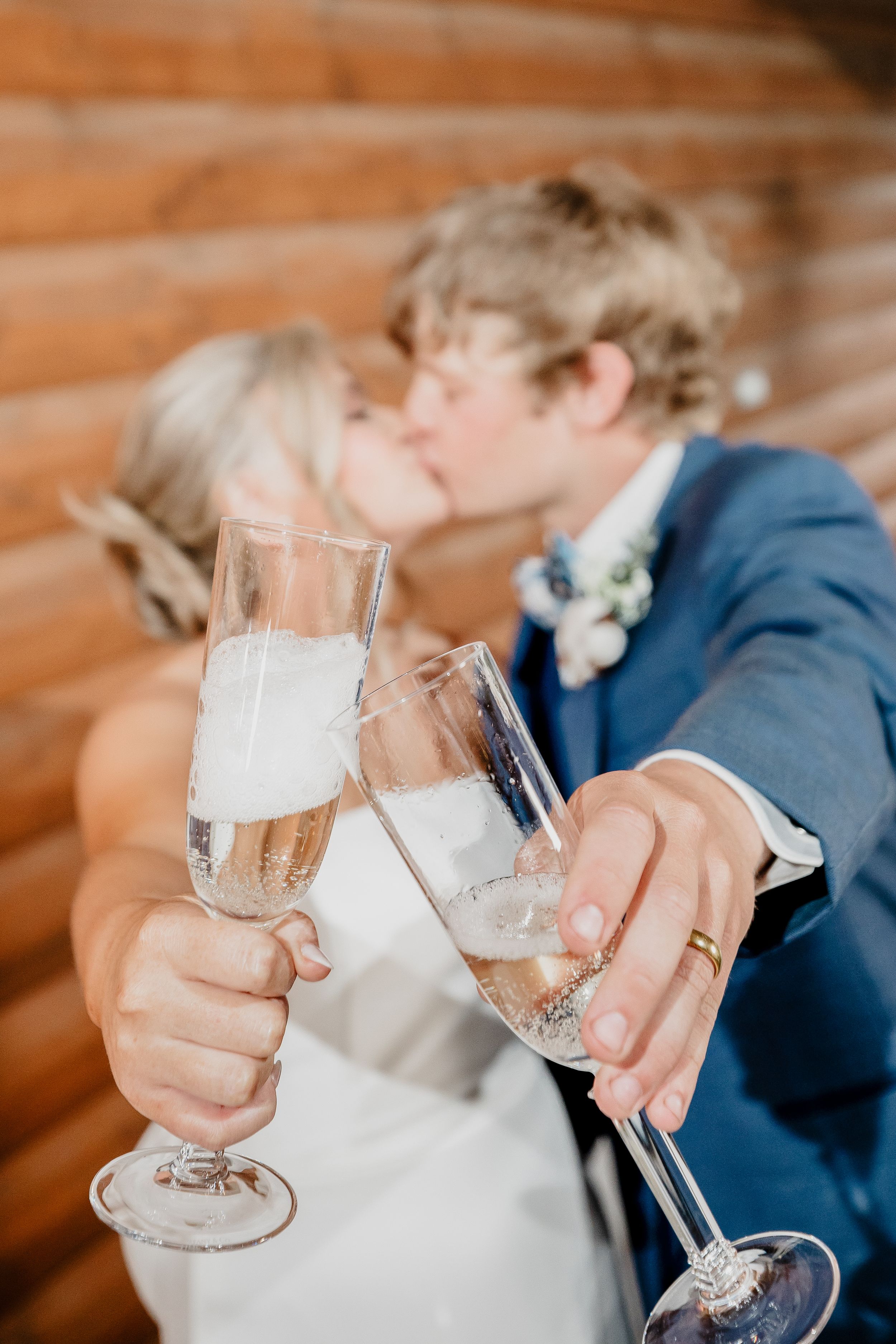 Newlywed couple kissing while clinking champagne flutes, wearing wedding attire near a rustic log wall.