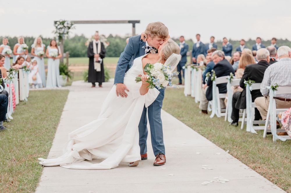 Bride and groom share a romantic dip kiss at outdoor wedding ceremony with guests seated along white aisle.