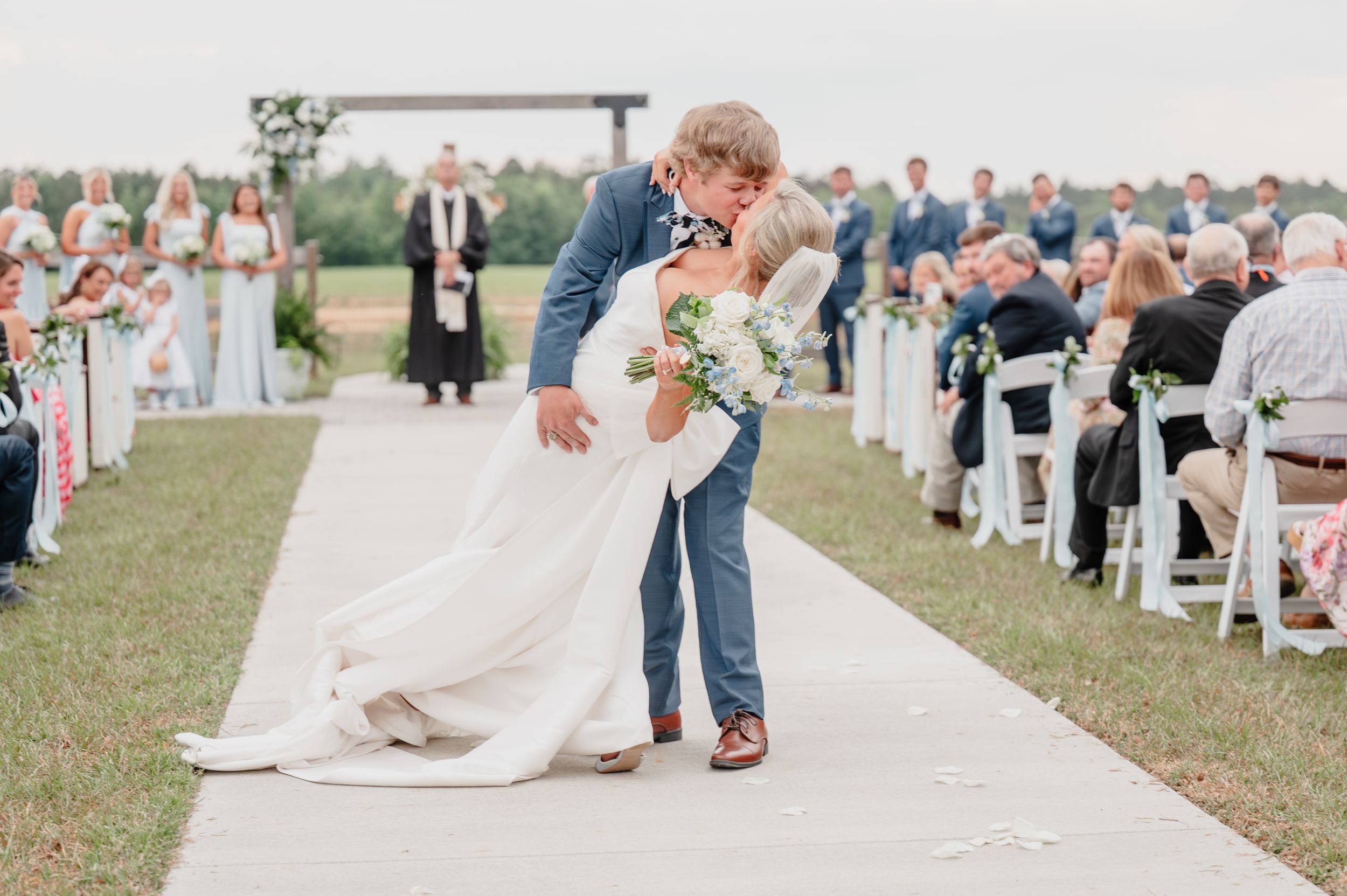 Bride and groom share a romantic dip kiss at outdoor wedding ceremony with guests seated along white aisle.