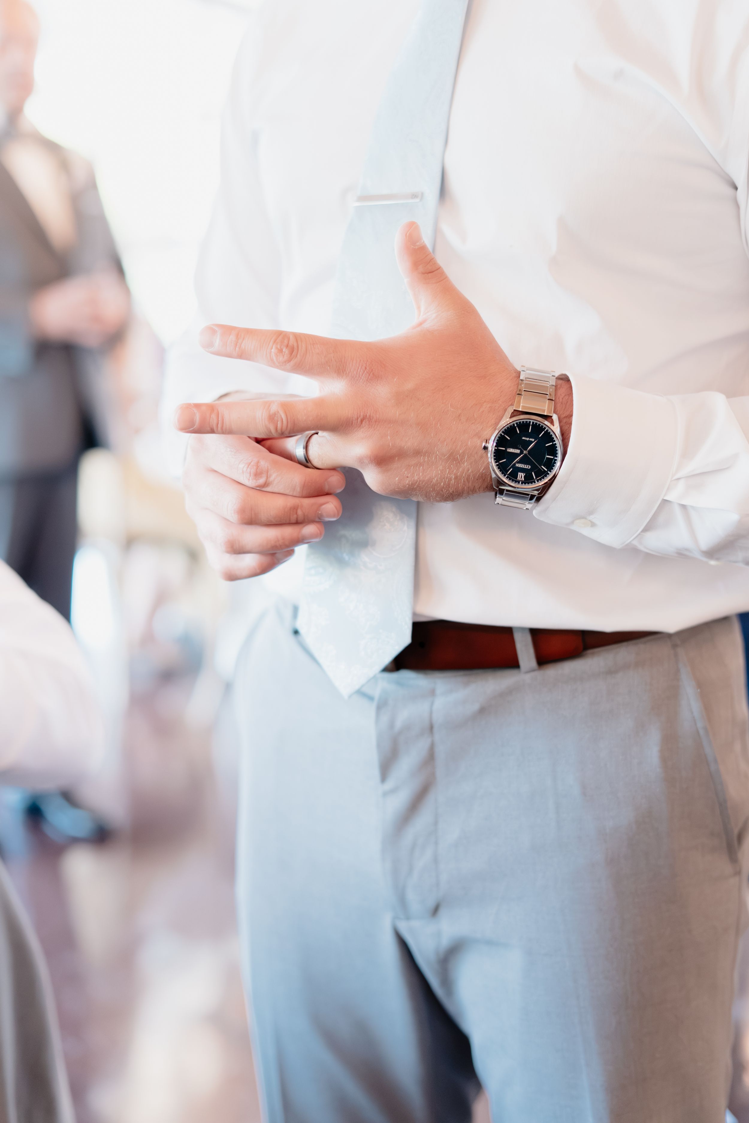 Man in white shirt and gray trousers adjusting light blue tie, wearing silver watch and ring.