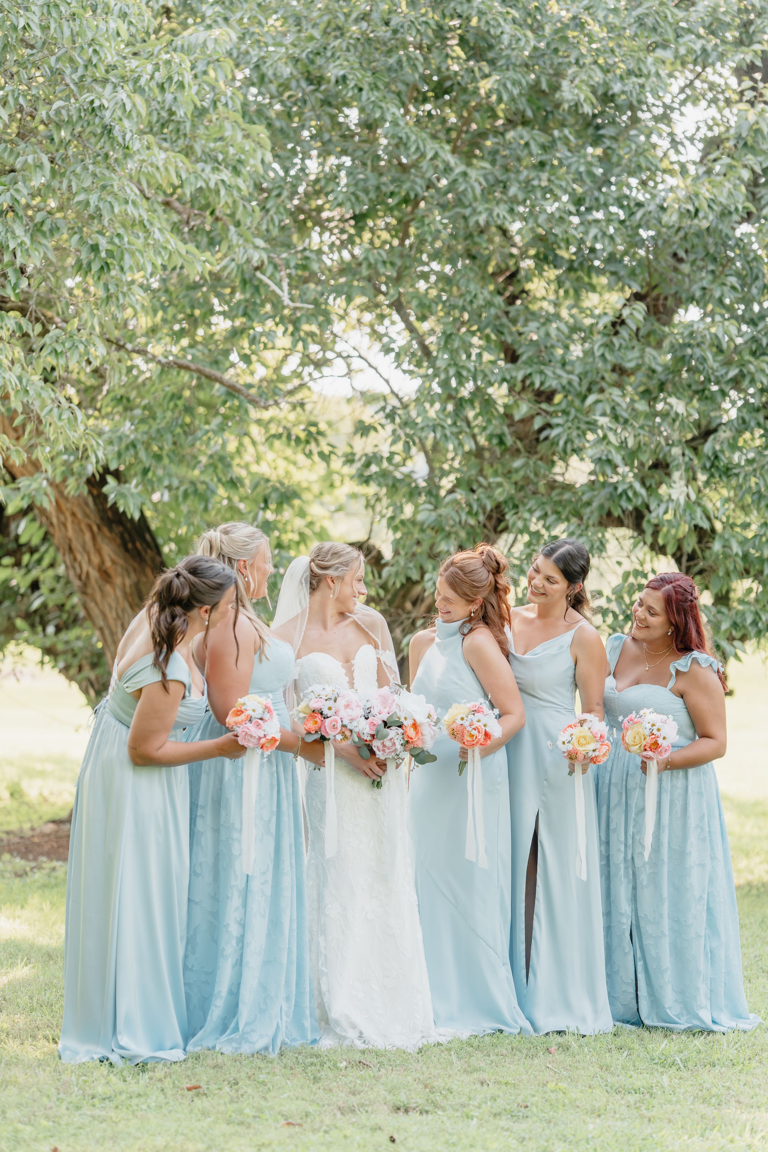 Bride in white gown with five bridesmaids in light blue dresses holding coral bouquets outdoors under a lush green tree.