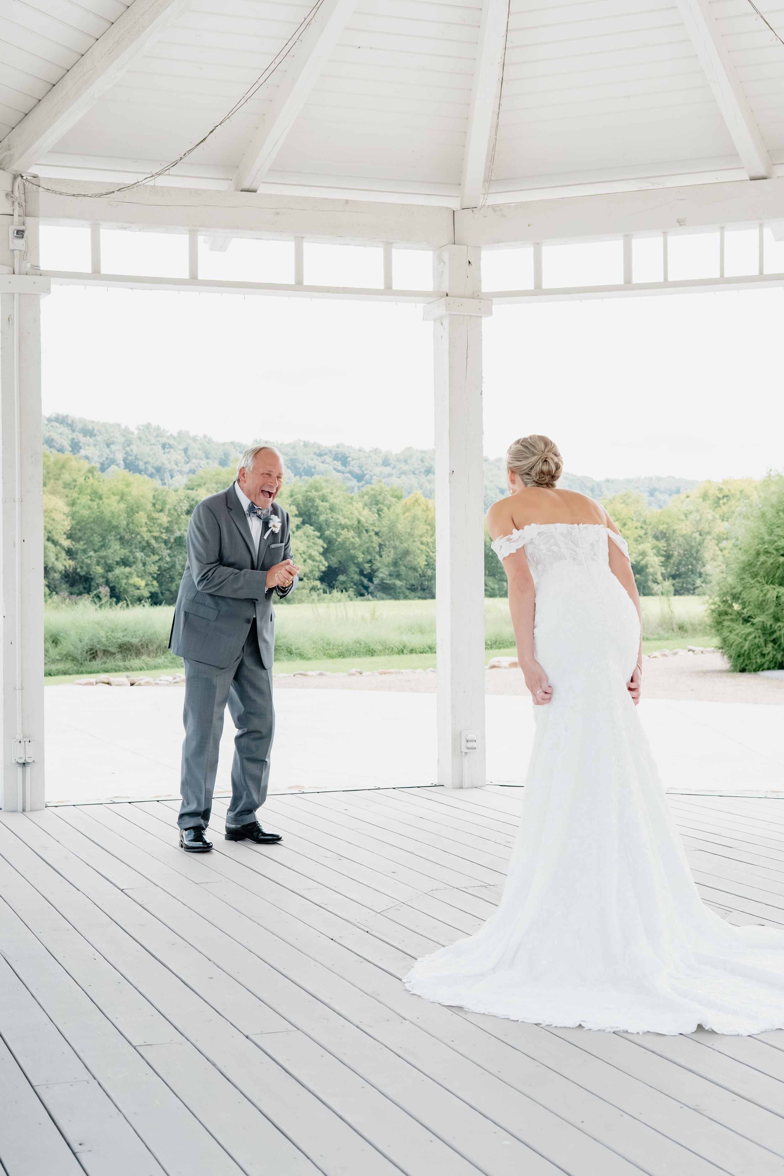 A bride in an off-shoulder white gown faces a tearful groom in a gray suit inside a white gazebo.