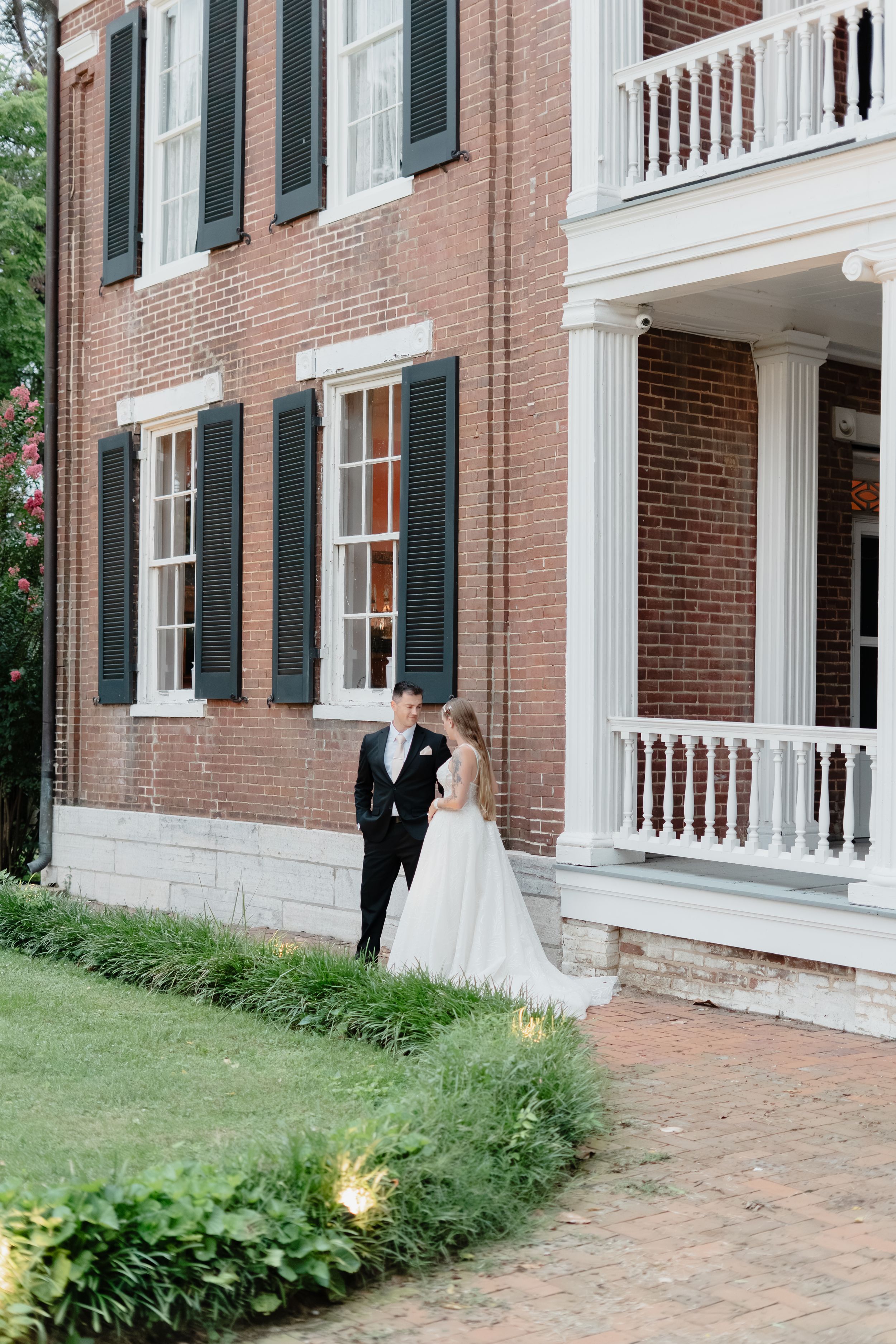 Bride and groom stand together outside a historic brick mansion with white columns and black shutters on their wedding day.