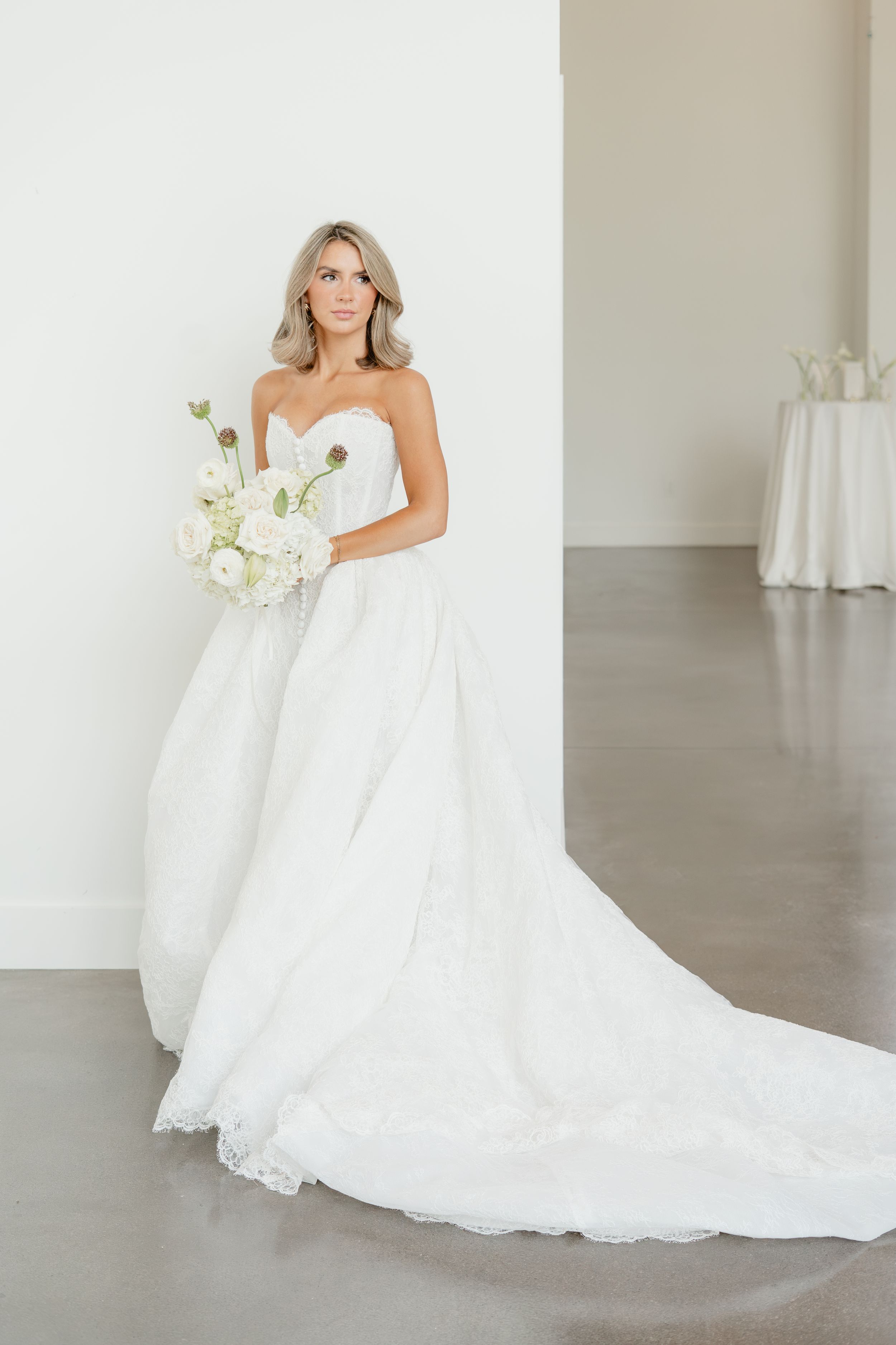 Bride in strapless white ball gown with lace train holds white floral bouquet in minimalist studio.