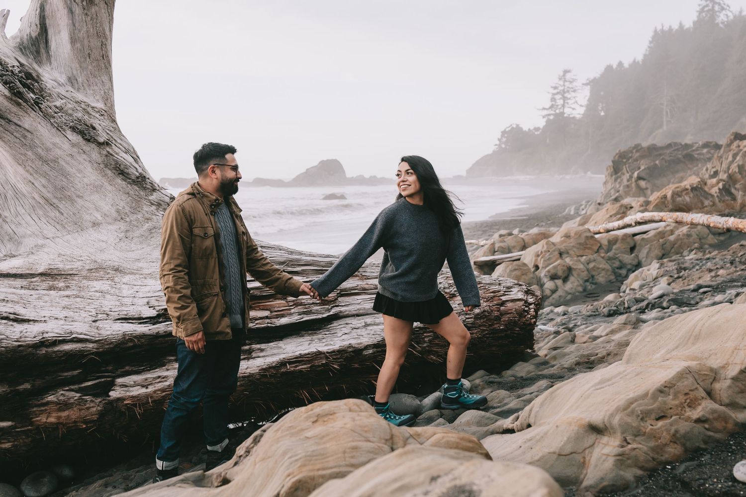 A couple is walking along the driftwood on a beach she is leading him and looking back at him.