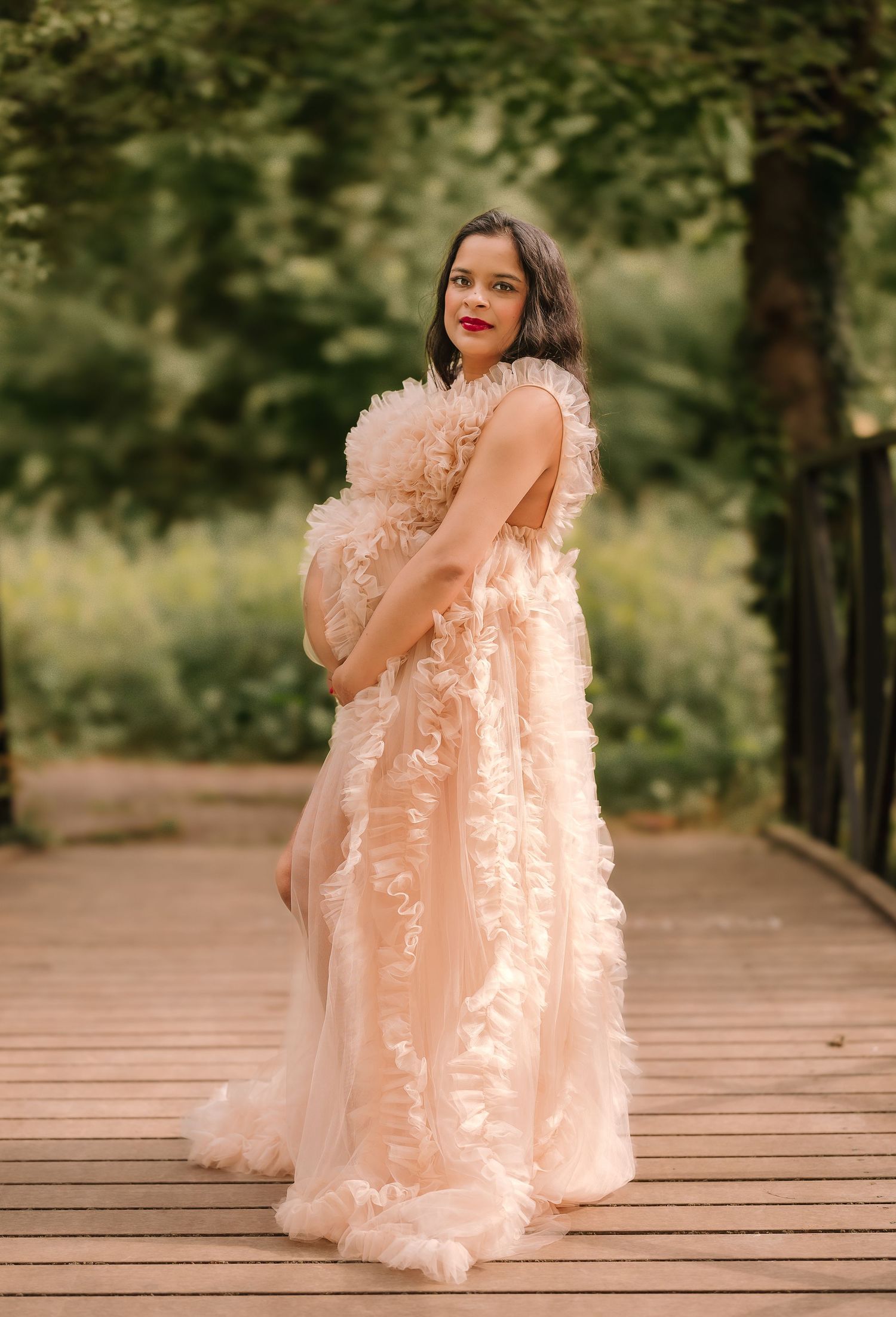 An expectant mother in a flowing peach gown poses on a wooden boardwalk surrounded by greenery.